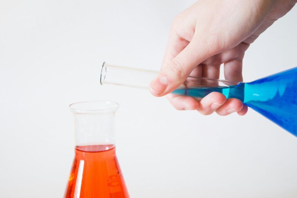 Hand pouring blue liquid from a test tube into a flask containing orange liquid.