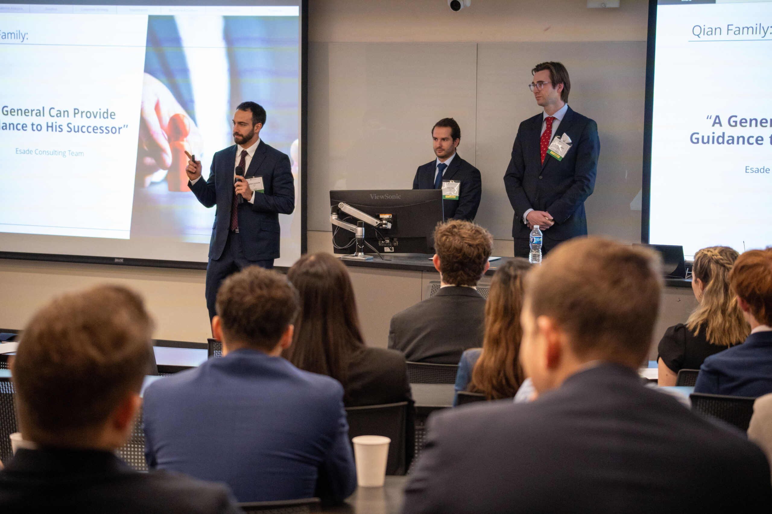 three students in suits stand in front of an audience presenting during a case competition