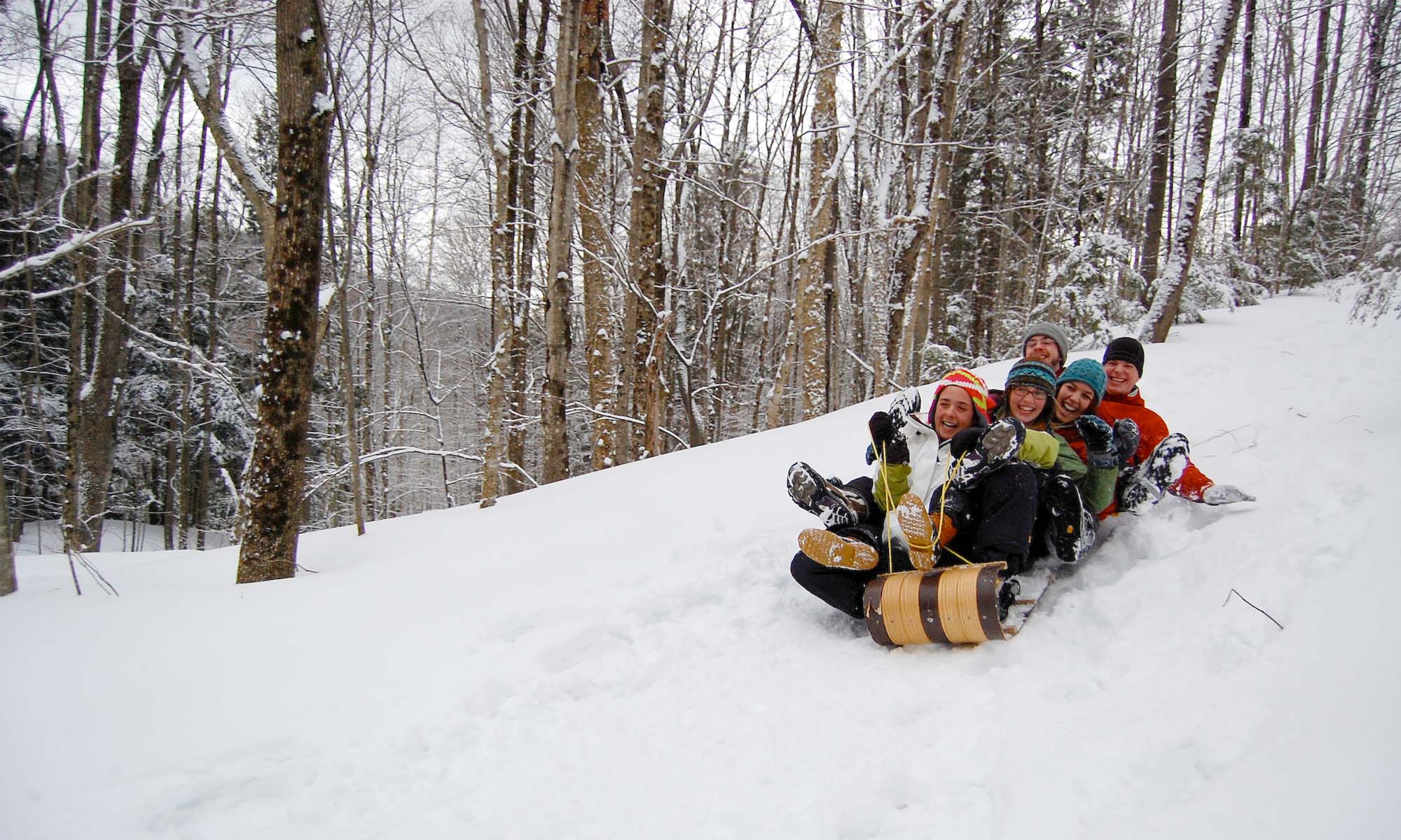 A group of female and male college students are riding on a toboggan down a snow covered trail in a forest.