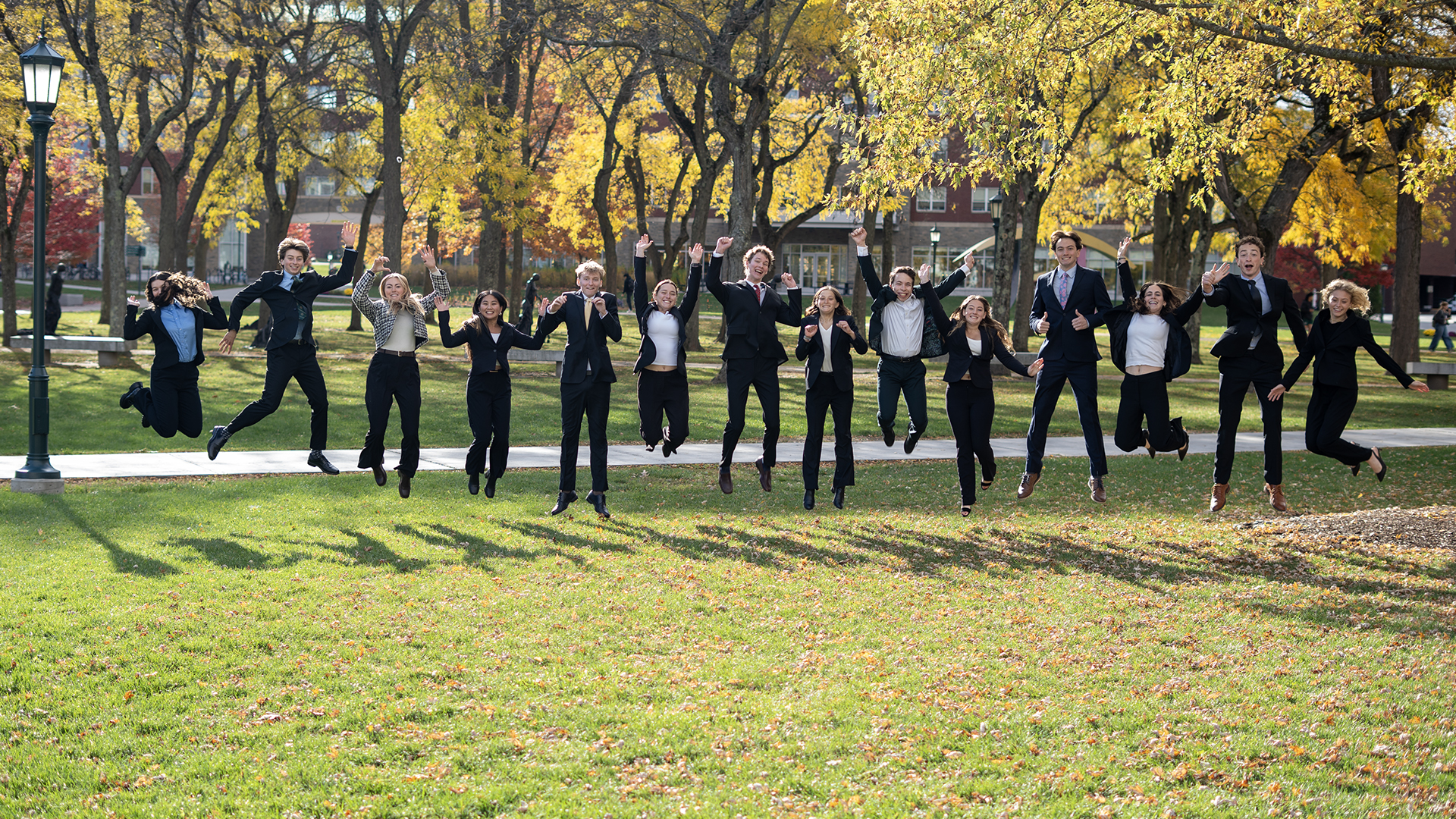 a line of students wearing business suits are caught jumping midair outside