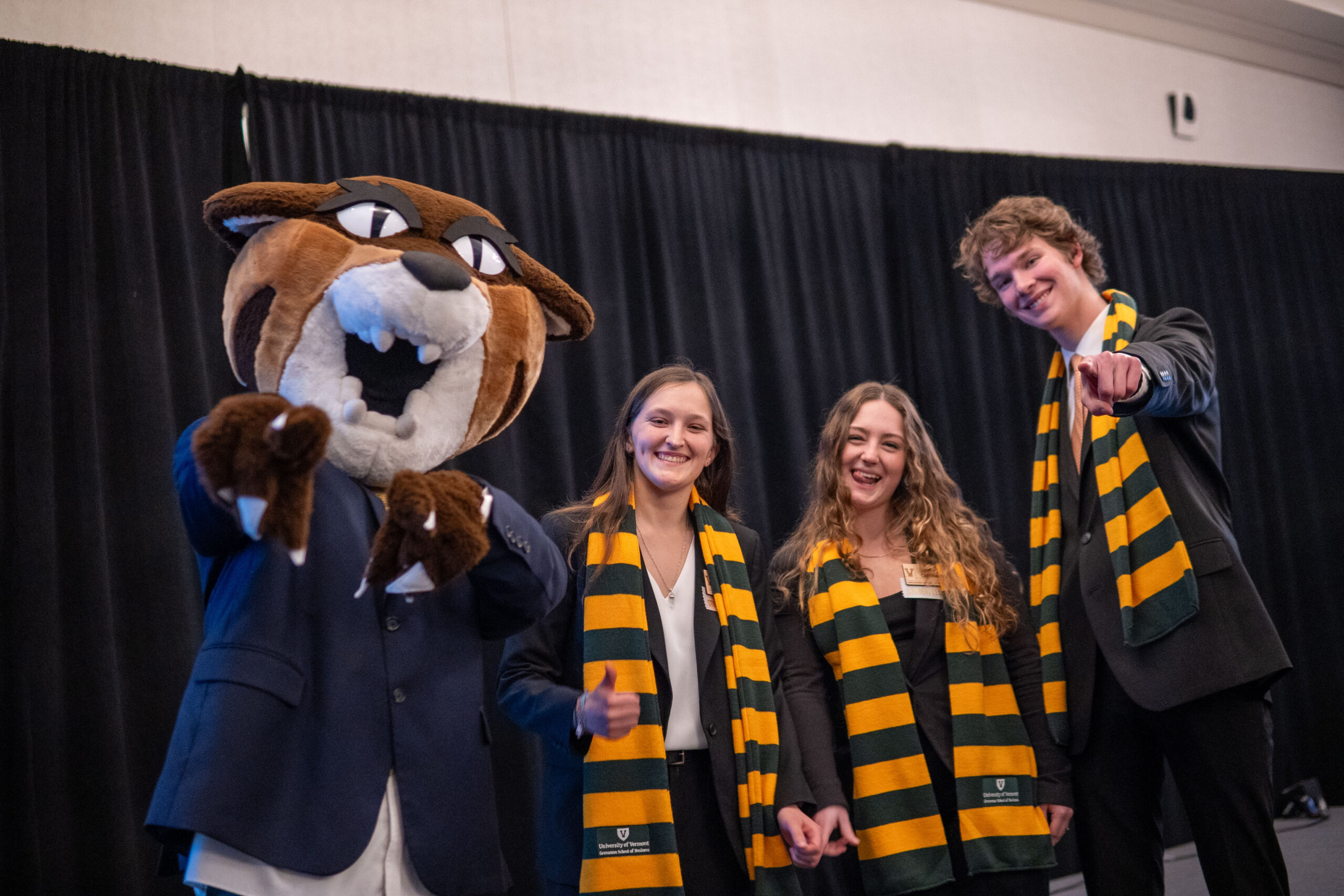 Rally Cat in a business jacket pointing at the camera with three business students wearing suits and UVM scarves.